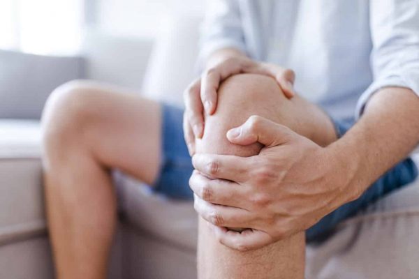 Man suffering from knee pain sitting sofa. A mature man massaging his painful knee. Man suffering from knee pain at home, closeup. Pain knee